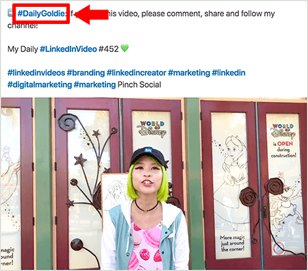 To jest zrzut ekranu, który ilustruje, jak Goldie Chan używa hashtagów w tekście swoich postów wideo na LinkedIn. Czerwone objaśnienia wskazują hashtag #DailyGoldie w tekście, który jest charakterystyczny dla jej postów wideo i pomaga jej śledzić udostępnienia. Post zawiera również inne trafne hashtagi, które pomagają ludziom znaleźć jej film, w tym #LinkedInVideo. Na obrazie wideo Goldie stoi przed kilkoma drzwiami na wystawie World of Disney. Jest Azjatką o zielonych włosach. Ma na sobie czarną czapkę LinkedIn, czarny naszyjnik choker, różową koszulę w makaronik oraz niebiesko-białą kurtkę.