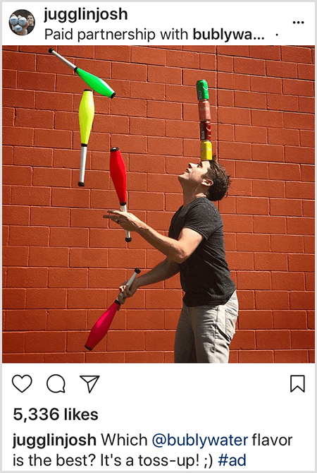 Josh Horton żongluje maczugami, które pasują do czterech puszek z wodą Bubly ustawionych na jego głowie. Tło stanowi mur z czerwonej cegły. Zdjęcie jest reklamą stworzoną przez Josha dla wody Bubly i zamieszczoną na swoim kanale na Instagramie z opisem, który smak wody Bubly jest najlepszy? To rzut do góry!